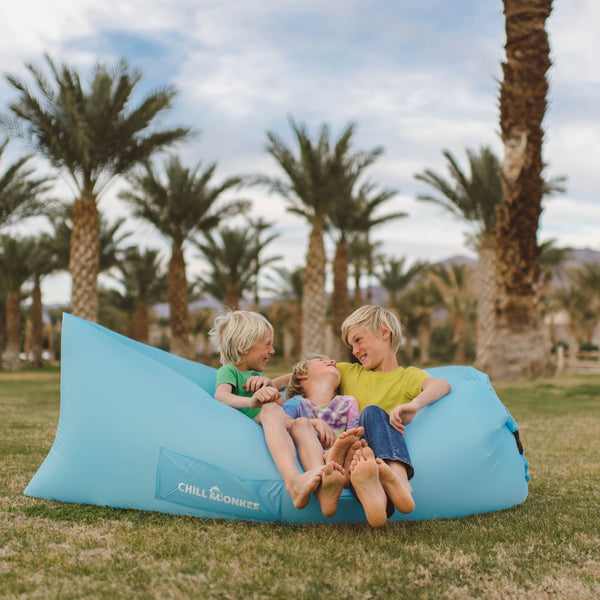 Three kids lounging on a Chill Monkee Sky Blue Lazy Lounger at a park under a palm tree.